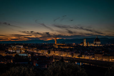 Illuminated bridge over river by buildings against sky at sunset