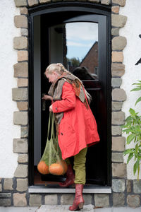 A middle-aged woman in a red cloak with a net of orange pumpkins opens the door