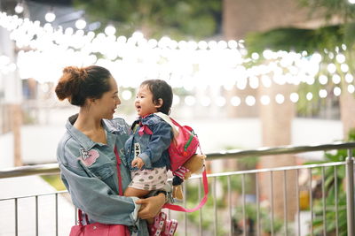 Mother carrying baby girl while standing against railing outdoors