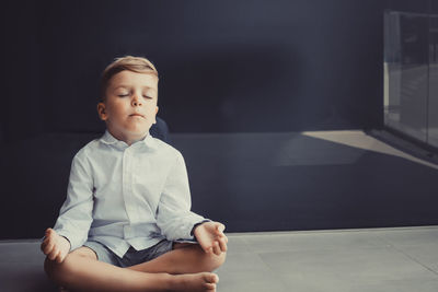 Full length of cute boy meditating at balcony against black wall