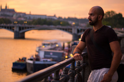 Thoughtful bald man standing by railing in city during sunset
