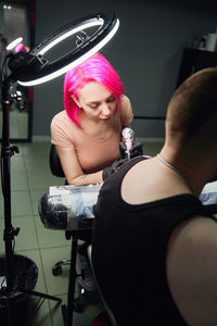 Serious tattoo master with pink hair in gloves using professional tattoo machine while making tattoo on shoulder of client in modern tattoo salon
