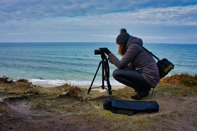 Side view of woman photographing sea while crouching at beach against sky