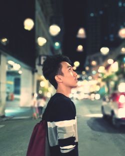Side view of young man looking up while standing on street in city at night