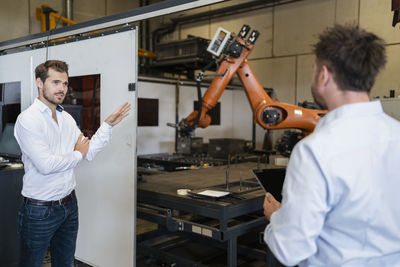 Businessman pointing at robotic arm machine while standing with colleague at factory