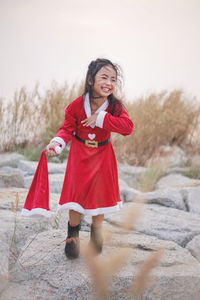 Cute girl wearing red dress holding santa hat while standing on rocks against sky