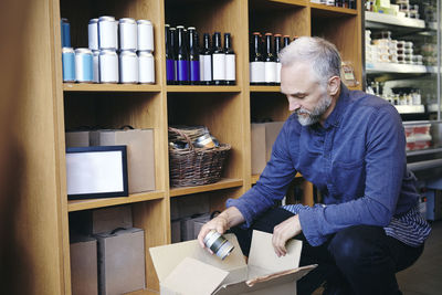 Mature salesman unpacking cardboard box by rack in deli