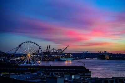 Illuminated ferris wheel by sea against sky at sunset