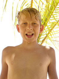 Portrait of cheerful shirtless wet boy standing against palm tree on sunny day