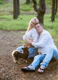 Full length of pregnant couple with dog against tree trunk