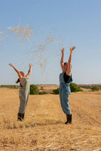 Side view of delighted female friends standing in dry field and throwing hay in air while having fun
