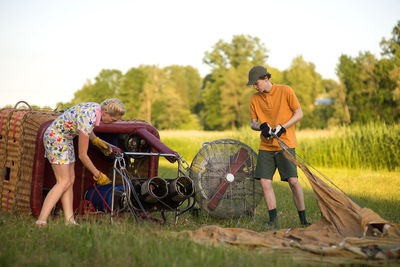 Boy assisting pilot in preparing hot air balloon on grassy field during sunset
