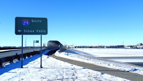 Signboard by road on snow covered field against clear blue sky