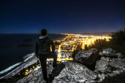 Rear view of man standing on rock at beach against sky at night