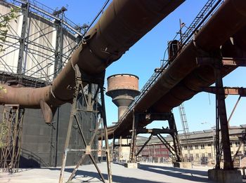 Low angle view of rusty pipes and water tower in decommissioned steel mill against clear blue sky