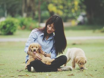 Full length of woman playing with puppy while sitting on grassy field at public park