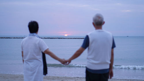 Rear view of couple holding hands while standing at beach against sky