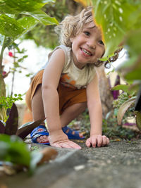 Small boy with curly hair smiling  crouched among the plants with his hands resting on the ground.