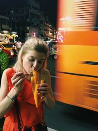 Young woman having fruit while standing on road in city at night