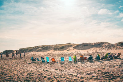 People relaxing on wooden chairs at beach against sky