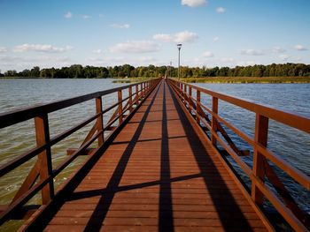 Diminishing perspective of footbridge over river against sky