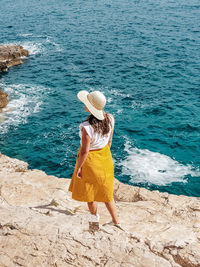 Young woman in yellow skirt standing on cliff above sea.