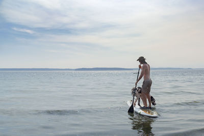 Shirtless man paddleboarding with dog in sea against sky