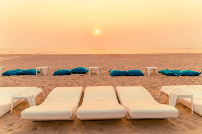 Lounge chairs on sand at beach against sky during sunset