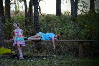 Full length of girl by her brother lying on wooden bench at forest