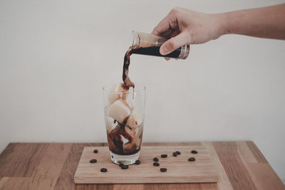 Cropped hand of person pouring drink in drinking glass on table against wall