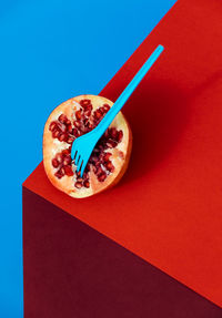 From above half of ripe pomegranate with seeds and pierced blue plastic fork placed on red table against blue background in studio