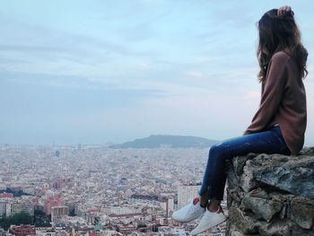 Side view of woman looking at city while sitting on cliff against sky