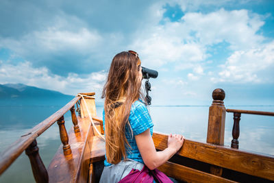 Side view of woman looking through binoculars while sitting in boat against sky