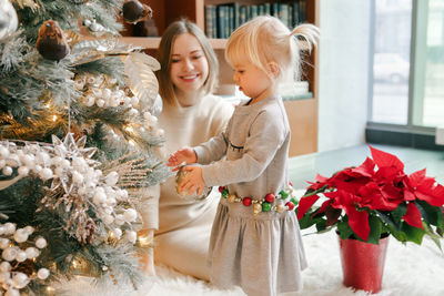 Happy caucasian mother with baby girl decorating christmas tree with ornaments baubles toy. 