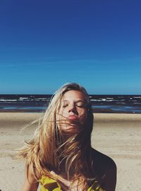 Young woman with eyes closed sticking out tongue while standing at beach against blue sky