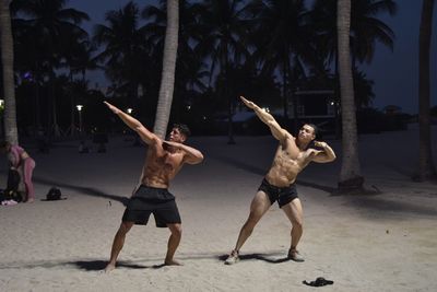 Full length of shirtless muscular men gesturing while standing at beach during night