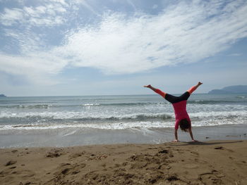 Rear view of woman doing handstand at beach against sky