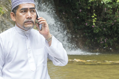 Thoughtful man talking on phone while sitting against waterfall