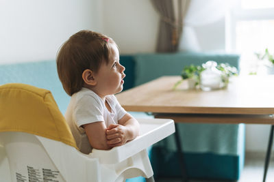 Charming little baby girl sitting in high chair for feeding at the bright kitchen at home