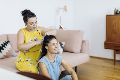 Cheerful woman braiding hair of friend in living room