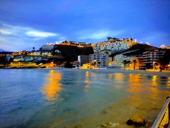 Illuminated buildings by sea against blue sky