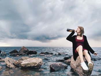 Carefree young woman drinking alcohol while sitting on rock against sea and cloudy sky