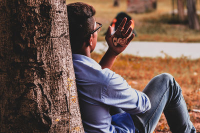 Rear view of young man sitting against tree trunk while looking at key ring