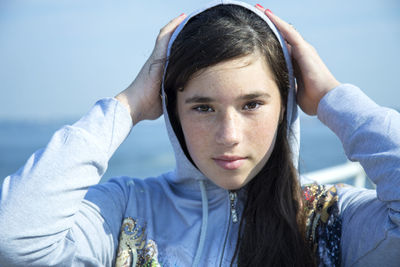 Close-up portrait of young woman in hooded shirt standing against sea