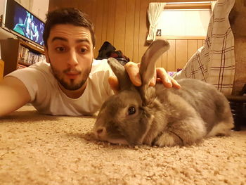 Young man patting rabbit on rug at home