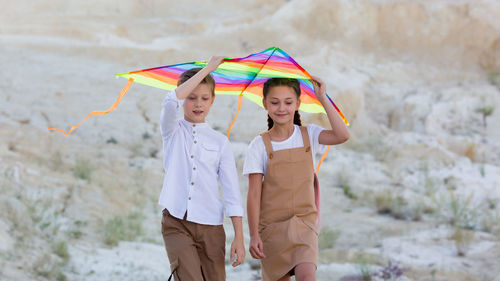 A girl and a boy are carrying bright kite with their hands raised high above their heads.