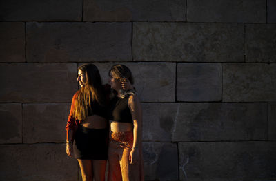 Two girls standing against stone wall during sunset