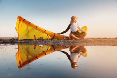 Attractive stylish young caucasian woman in cap sunglasses and kitesurfer outfit sits on the sandy