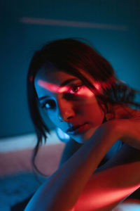 Tranquil young female model in dress sitting on floor and leaning on knee while looking at camera in dark studio with colorful lights