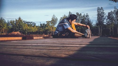 Side view of young man exercising while sitting on boardwalk against sky